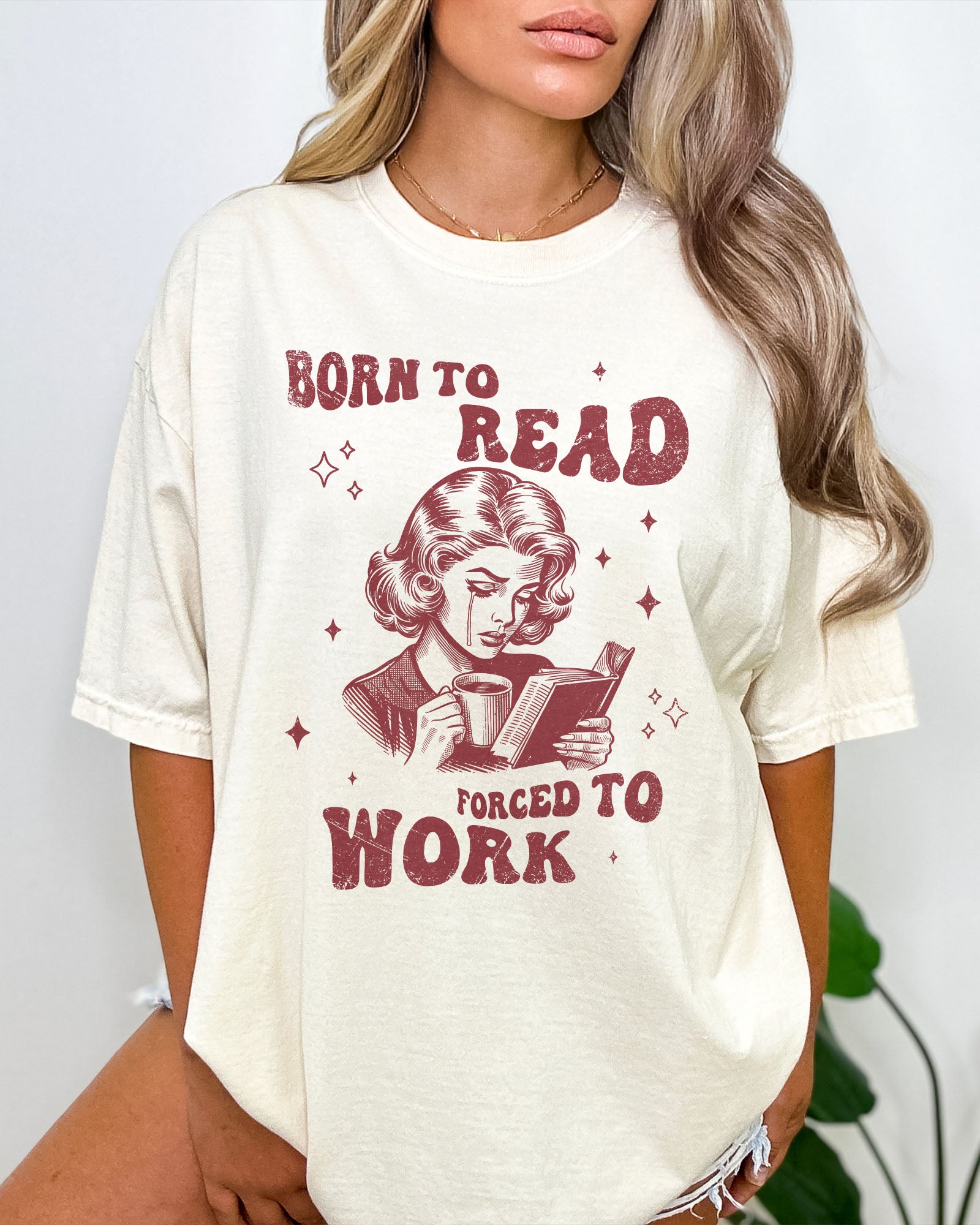 Woman wearing an ivory “Born To Read Forced To Work” shirt with a retro graphic of a sad woman holding a book.