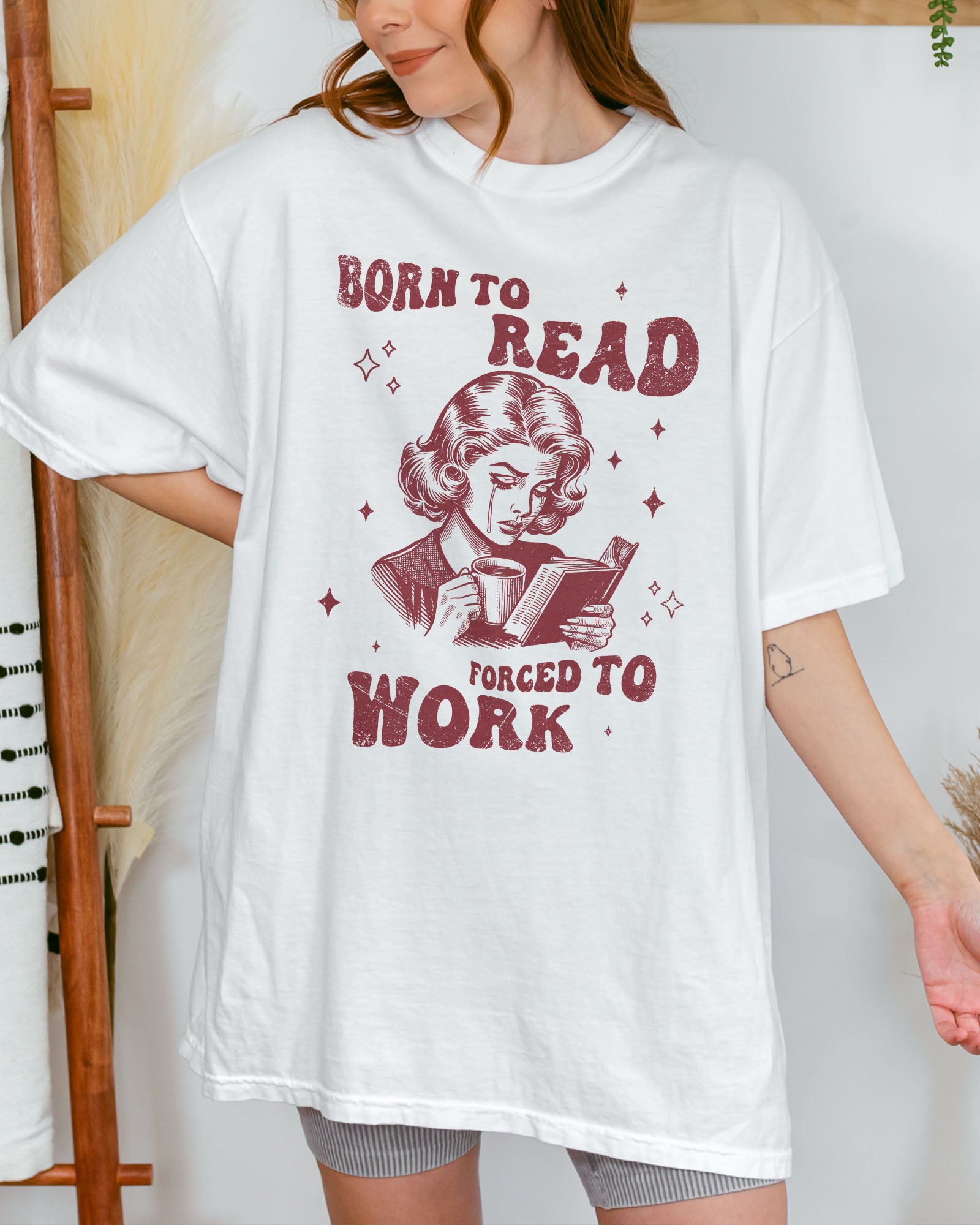 Woman wearing a white “Born To Read Forced To Work” shirt with a retro graphic of a sad woman holding a book.