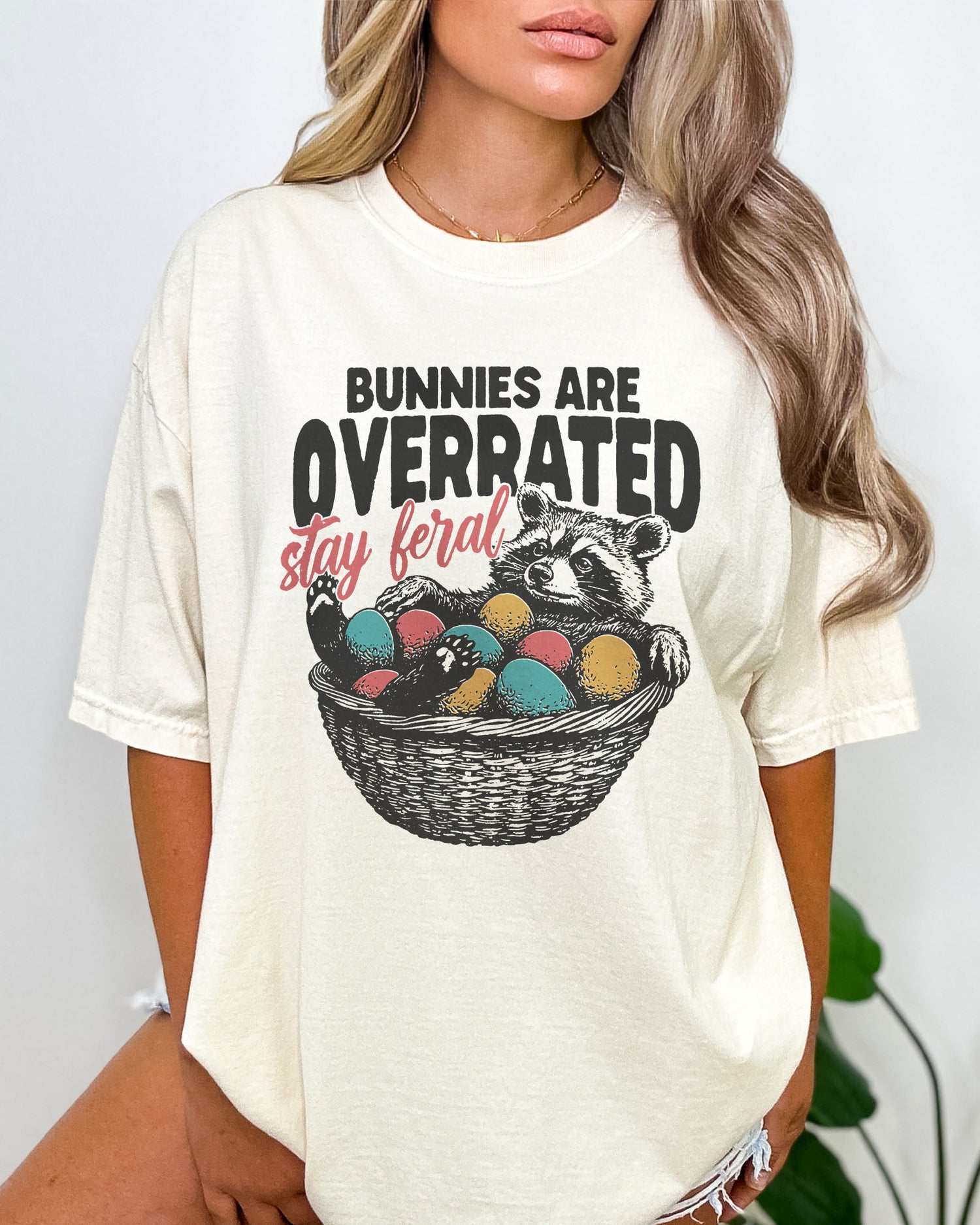 Model wearing natural color shirt with 'Bunnies Are Overrated - Stay Feral' easter raccoon graphic design