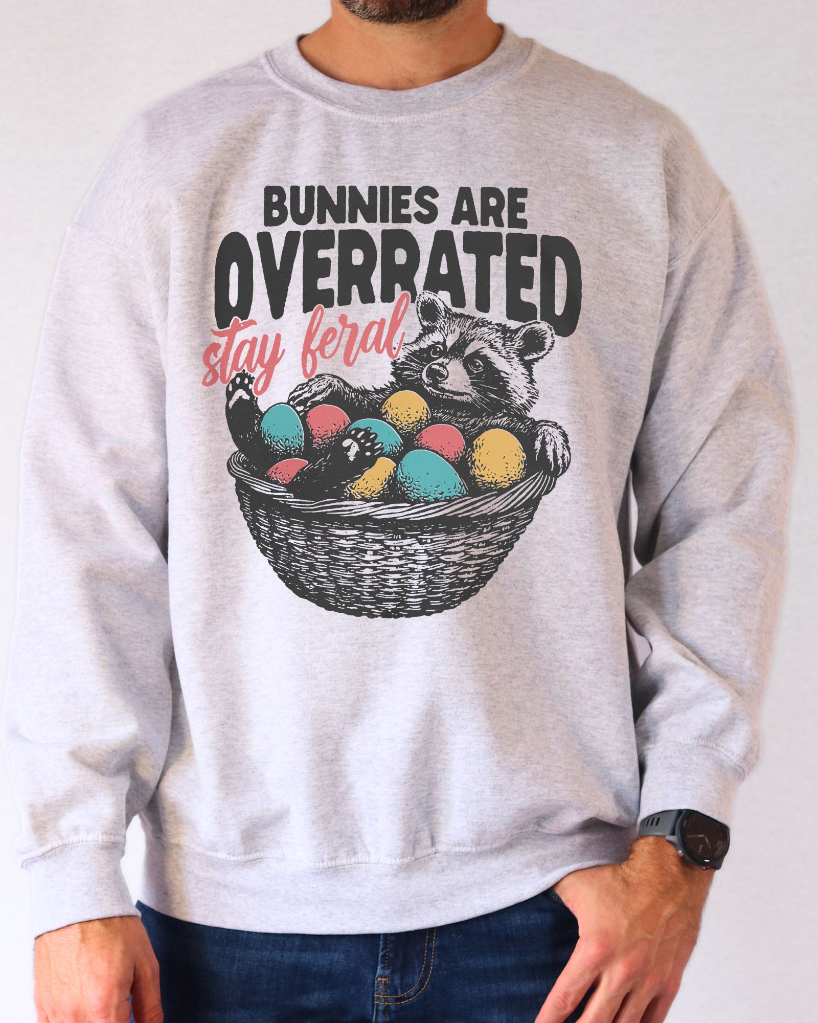 Man wearing an ash gray sweatshirt with a funny retro-style graphic of a raccoon in an Easter egg basket and the quote "Bunnies Are Overrated, Stay Feral".