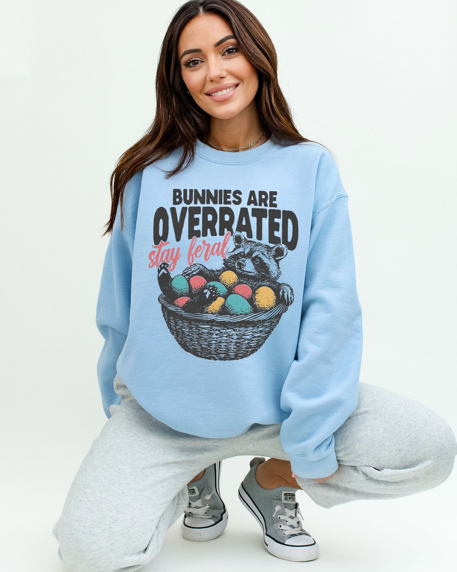 Woman wearing a light blue sweatshirt with a funny retro-style graphic of a raccoon in an Easter egg basket and the quote "Bunnies Are Overrated, Stay Feral".