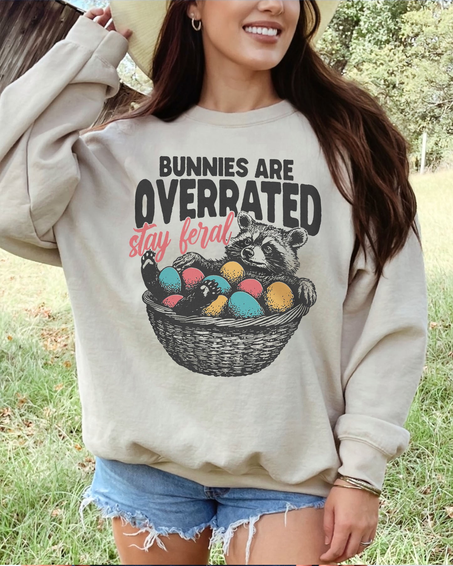 Woman wearing a sand beige sweatshirt with a funny retro-style graphic of a raccoon in an Easter egg basket and the quote "Bunnies Are Overrated, Stay Feral".