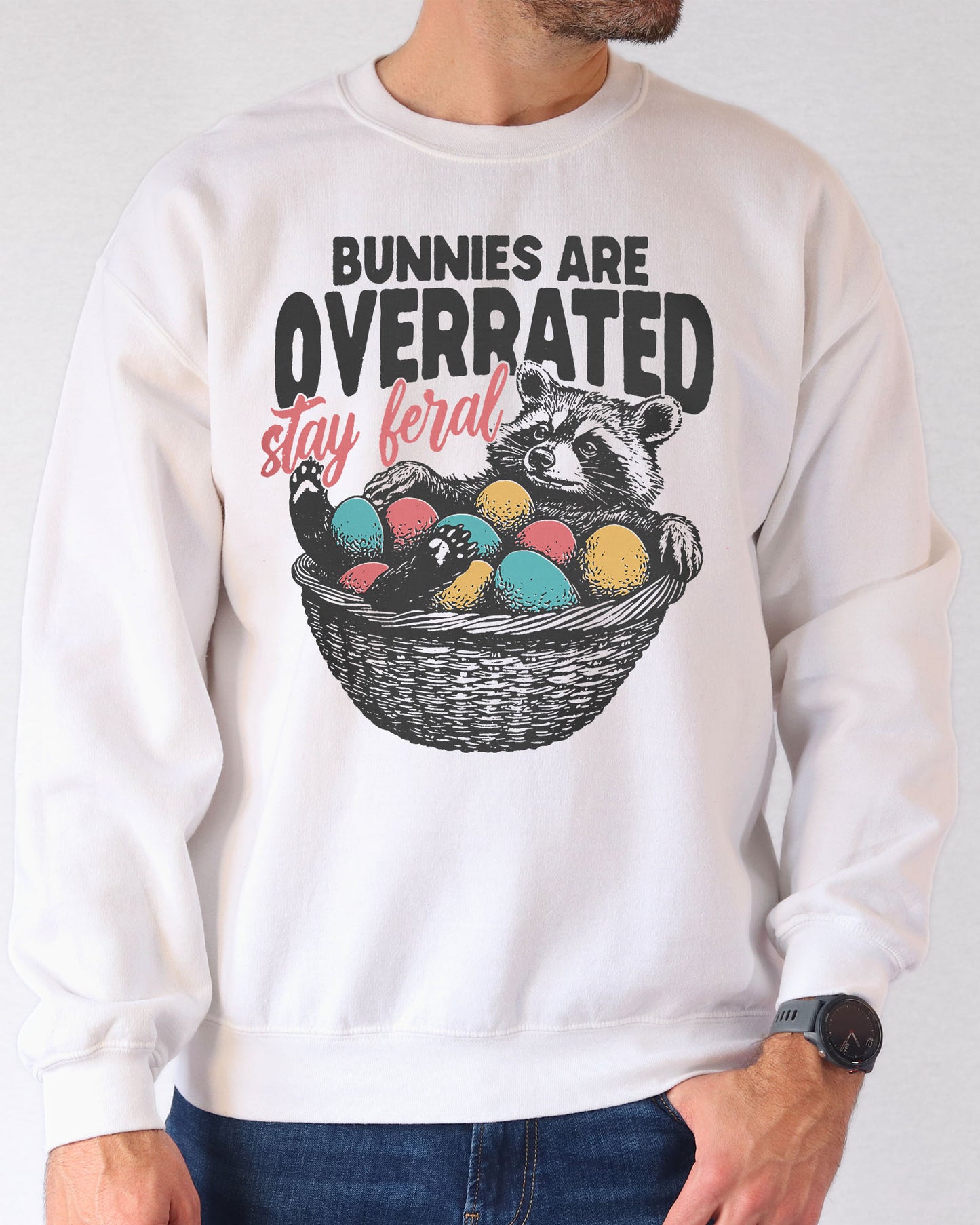 Man wearing a white sweatshirt with a funny retro-style graphic of a raccoon in an Easter egg basket and the quote "Bunnies Are Overrated, Stay Feral".