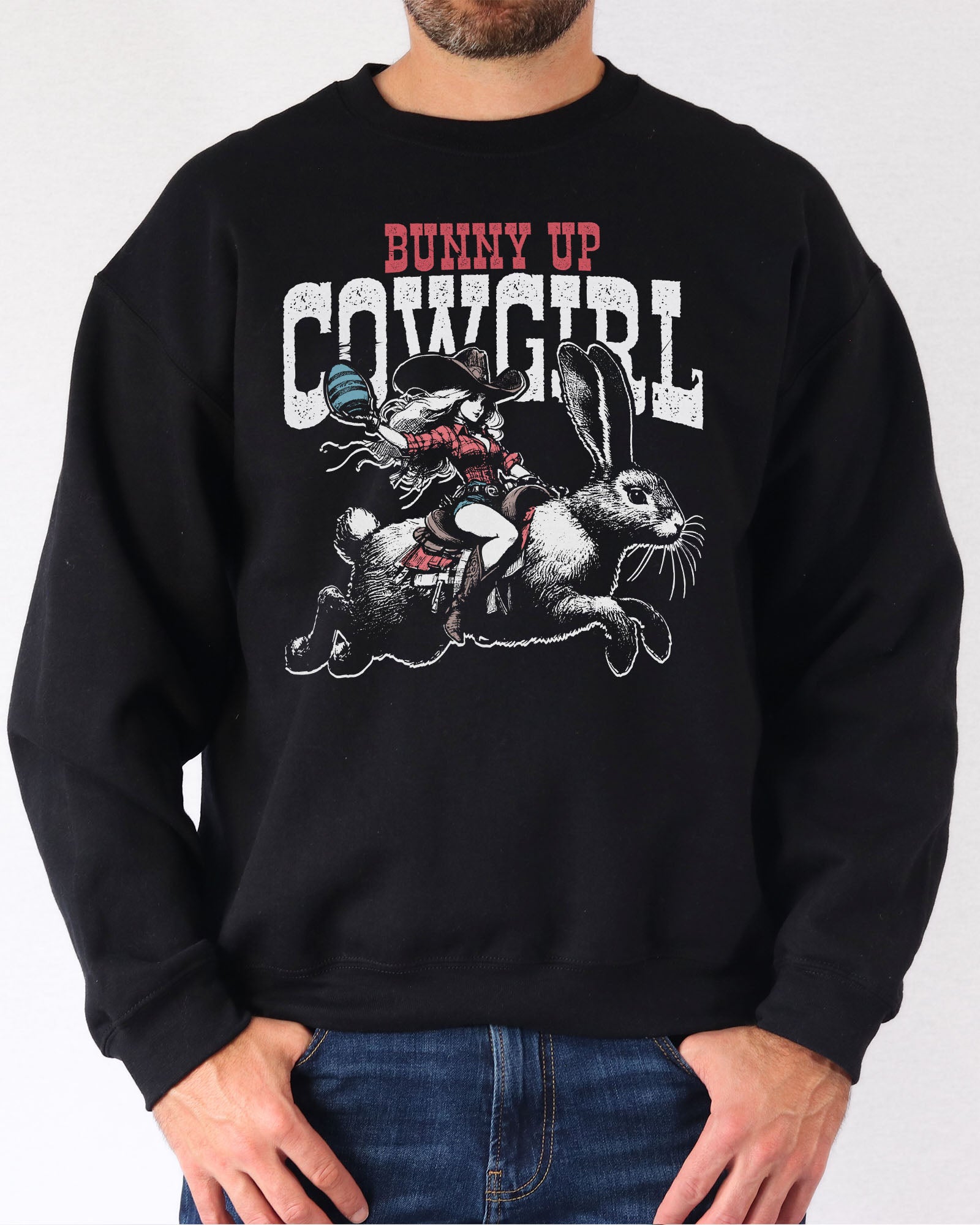 Man wearing a black sweatshirt featuring a funny retro graphic of a cowgirl riding an easter bunny and the quote 'Bunny Up Cowgirl'.