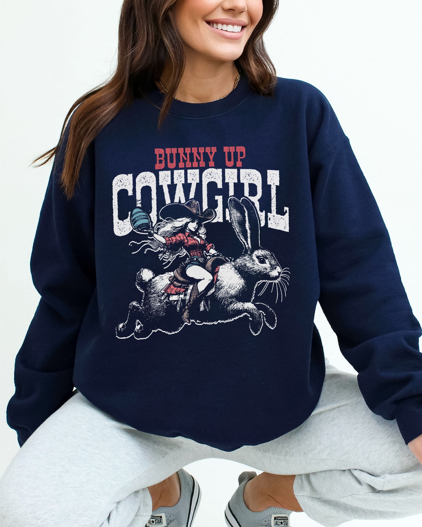Woman wearing a navy blue sweatshirt featuring a funny retro graphic of a cowgirl riding an easter bunny and the quote 'Bunny Up Cowgirl'.