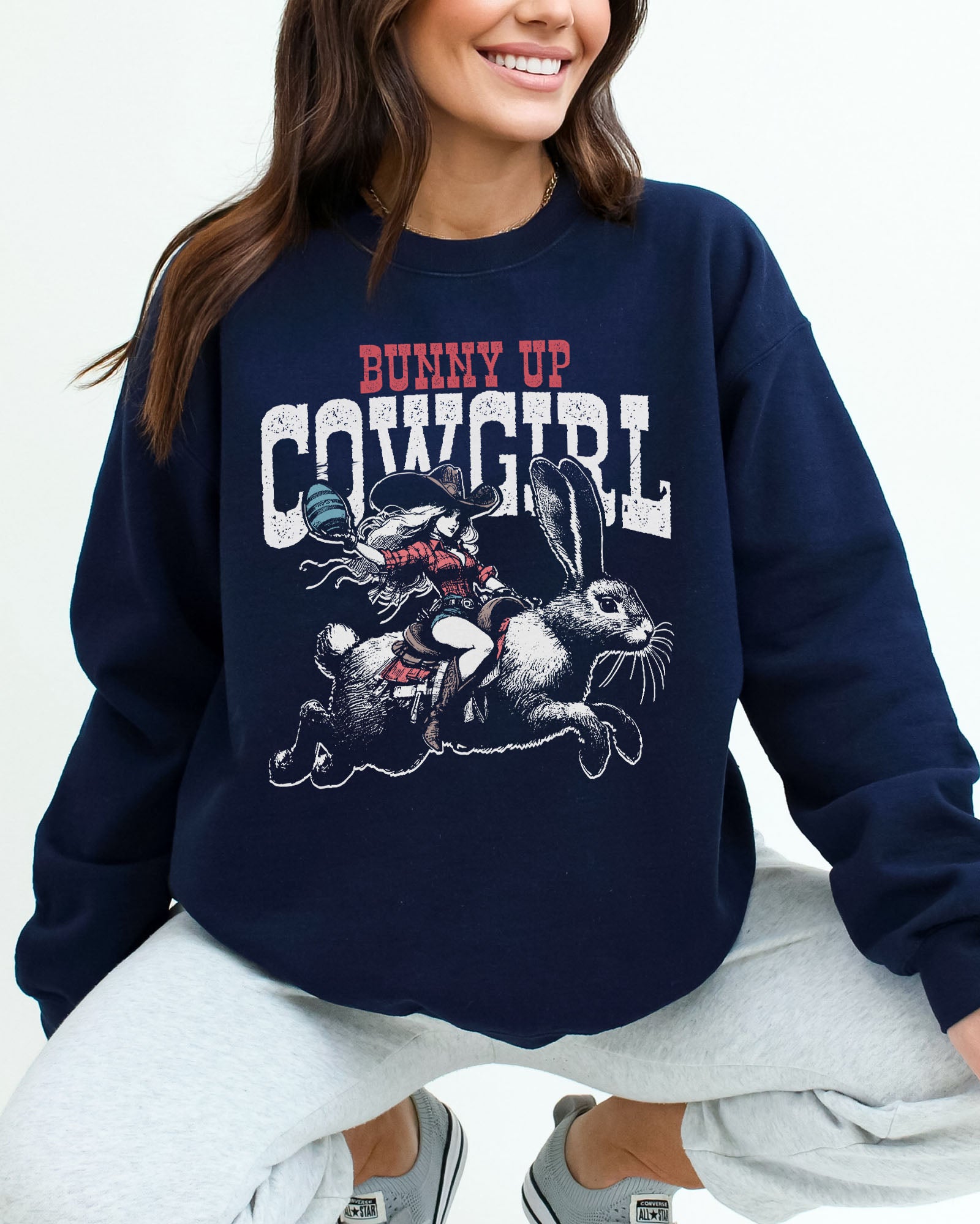 Woman wearing a navy blue sweatshirt featuring a funny retro graphic of a cowgirl riding an easter bunny and the quote 'Bunny Up Cowgirl'.