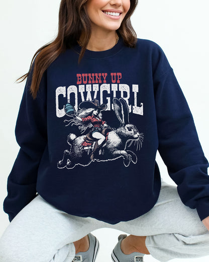 Woman wearing a navy blue sweatshirt featuring a funny retro graphic of a cowgirl riding an easter bunny and the quote 'Bunny Up Cowgirl'.