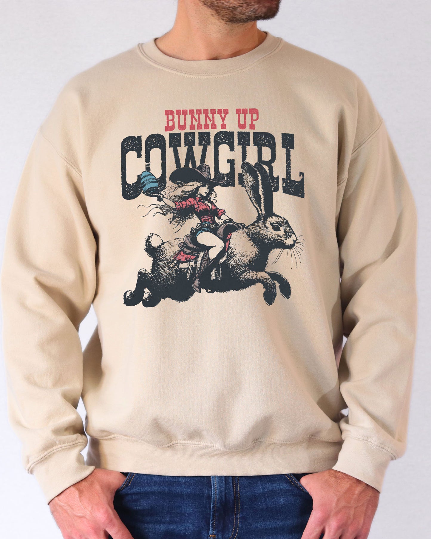 Man wearing a sand beige sweatshirt featuring a funny retro graphic of a cowgirl riding an easter bunny and the quote 'Bunny Up Cowgirl'.