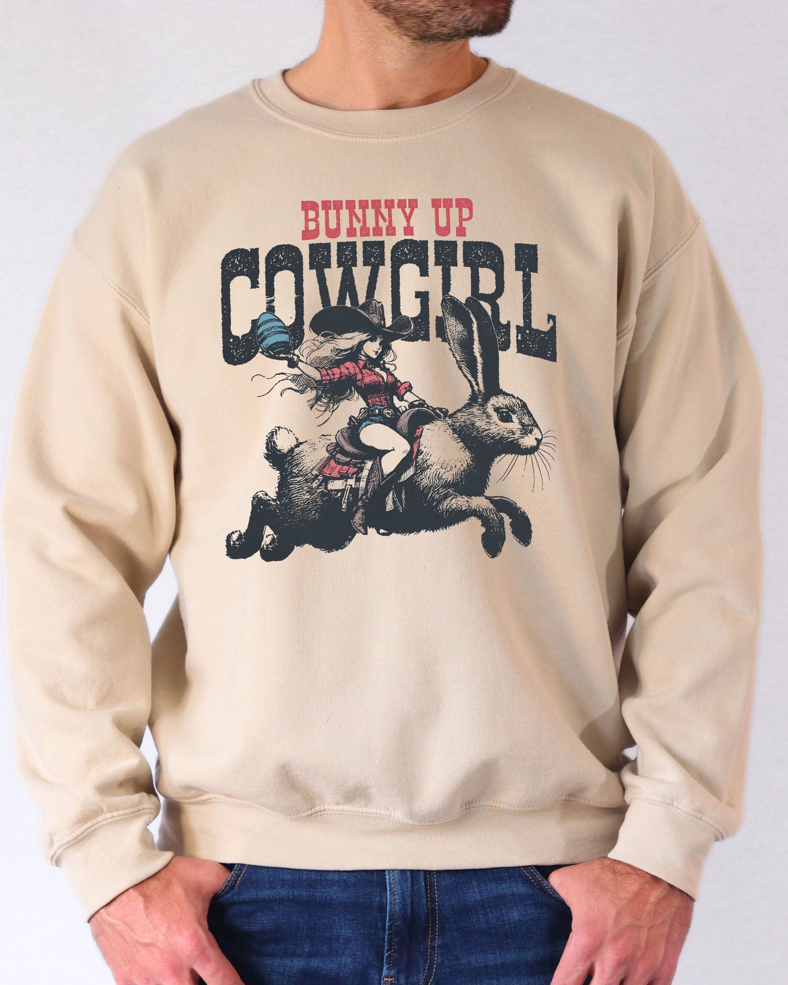 Man wearing a sand beige sweatshirt featuring a funny retro graphic of a cowgirl riding an easter bunny and the quote 'Bunny Up Cowgirl'.
