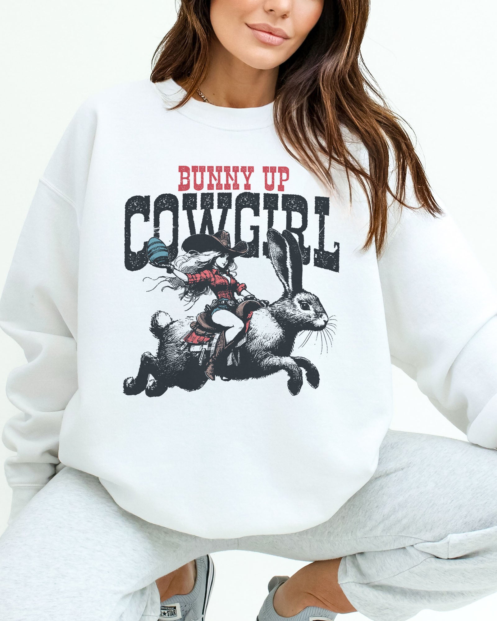 Woman wearing a white sweatshirt featuring a funny retro graphic of a cowgirl riding an easter bunny and the quote 'Bunny Up Cowgirl'.