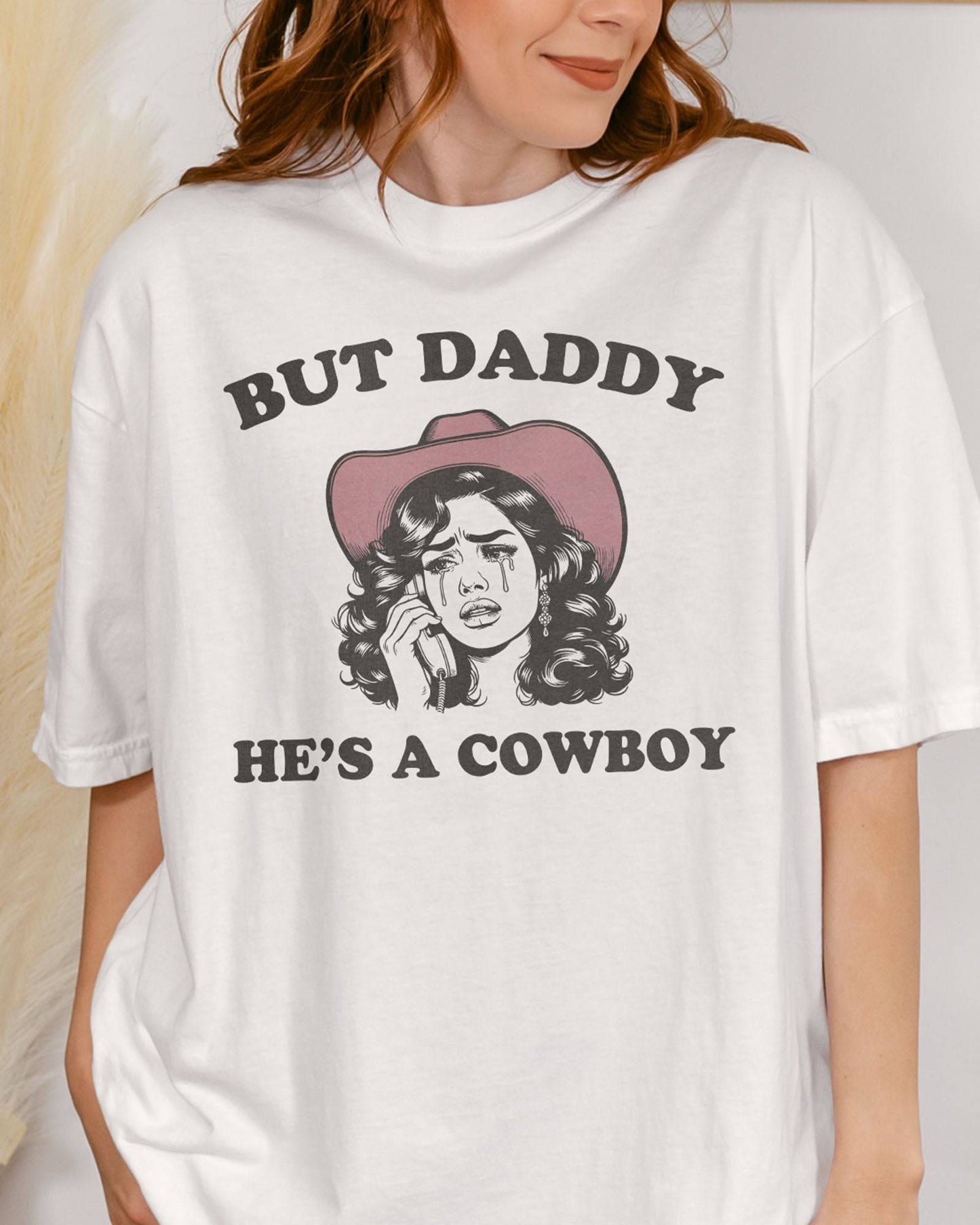 Close-up of woman wearing a white "But Daddy He's a Cowboy" shirt with a funny retro graphic of a cowgirl crying while talking on the phone.