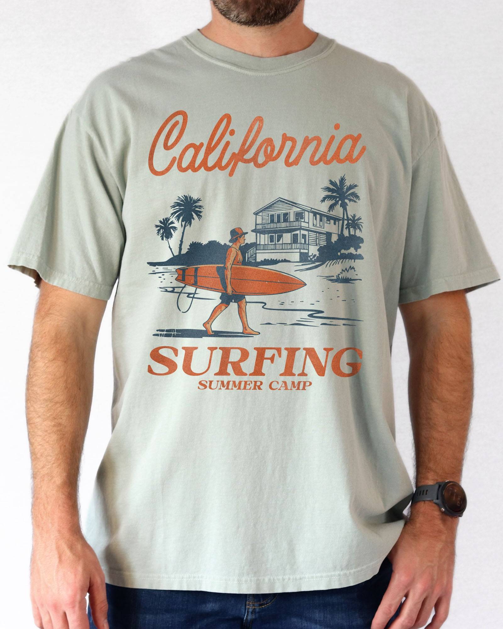 Man wearing a bay-colored "California Surfing Summer Camp" shirt with a retro graphic of a surfer carrying a surfboard past a beach house.