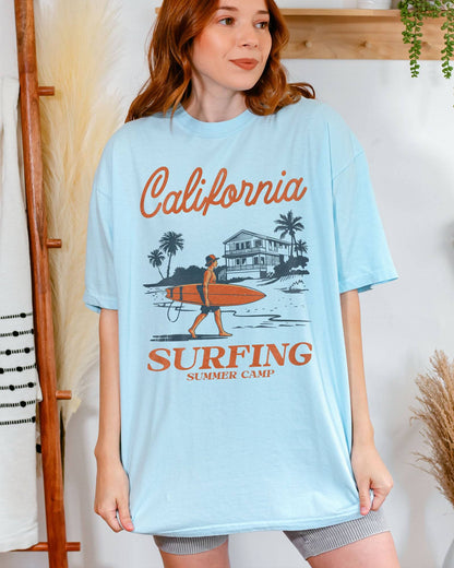 Woman wearing a chambray-colored "California Surfing Summer Camp" shirt with a retro graphic of a surfer carrying a surfboard past a beach house.