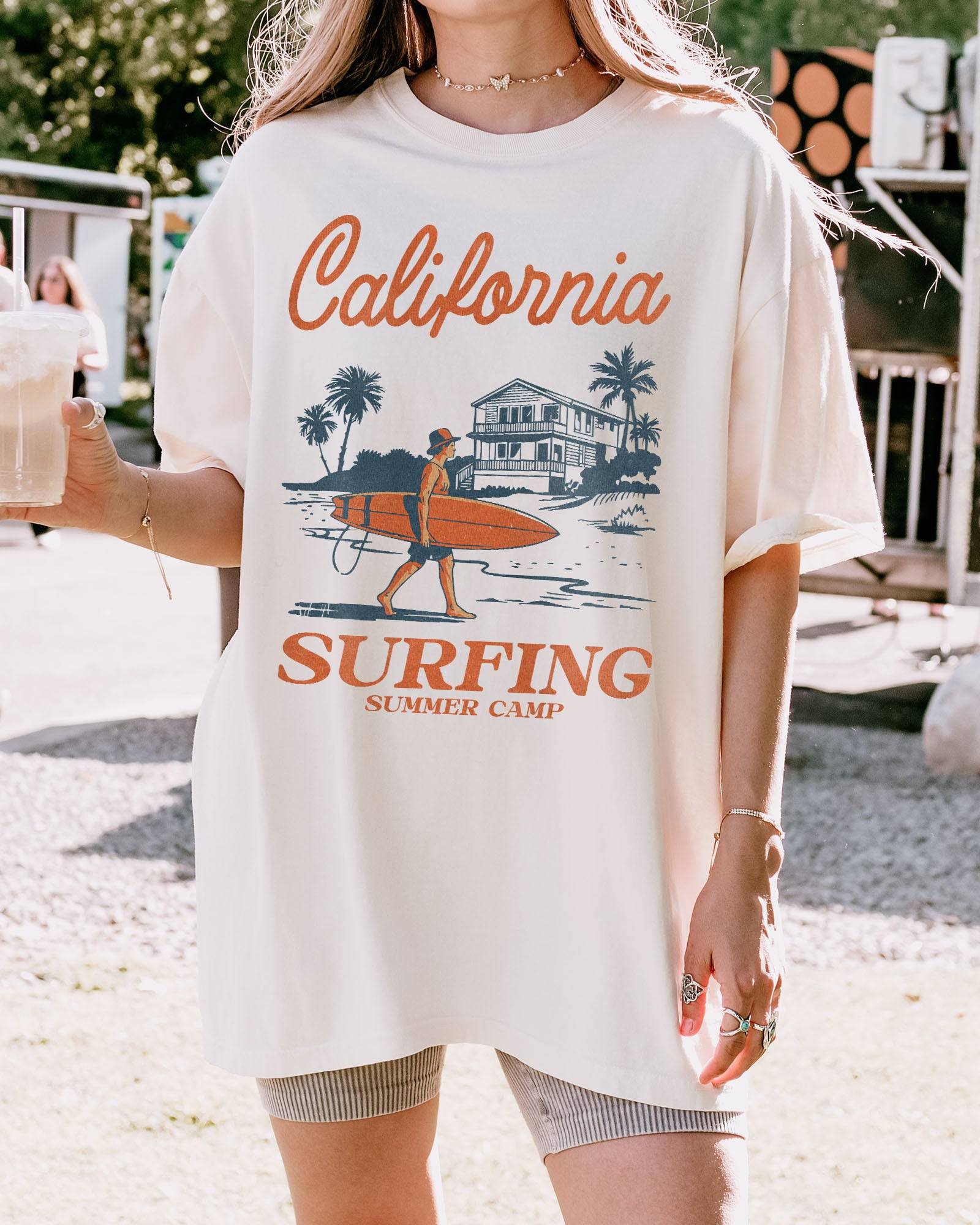 Woman wearing an ivory-colored "California Surfing Summer Camp" shirt with a retro graphic of a surfer carrying a surfboard past a beach house.