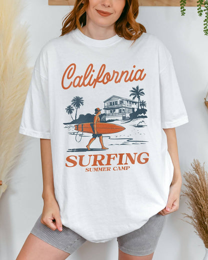Woman wearing a white-colored "California Surfing Summer Camp" shirt with a retro graphic of a surfer carrying a surfboard past a beach house.
