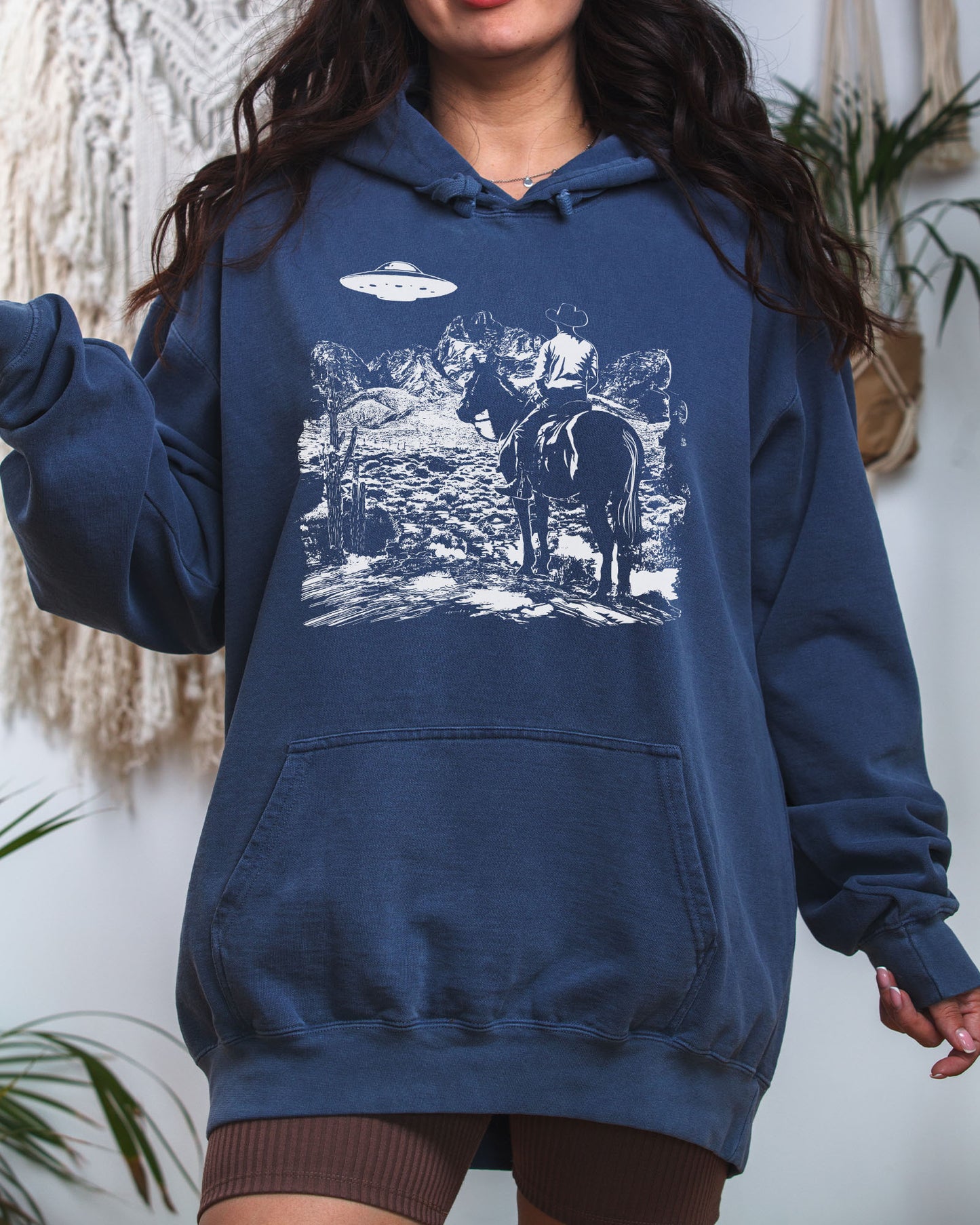 Woman wearing a denim-colored "Cowboy UFO Encounter" hoodie with a retro graphic of a cowboy riding through a desert as a UFO hovers above.