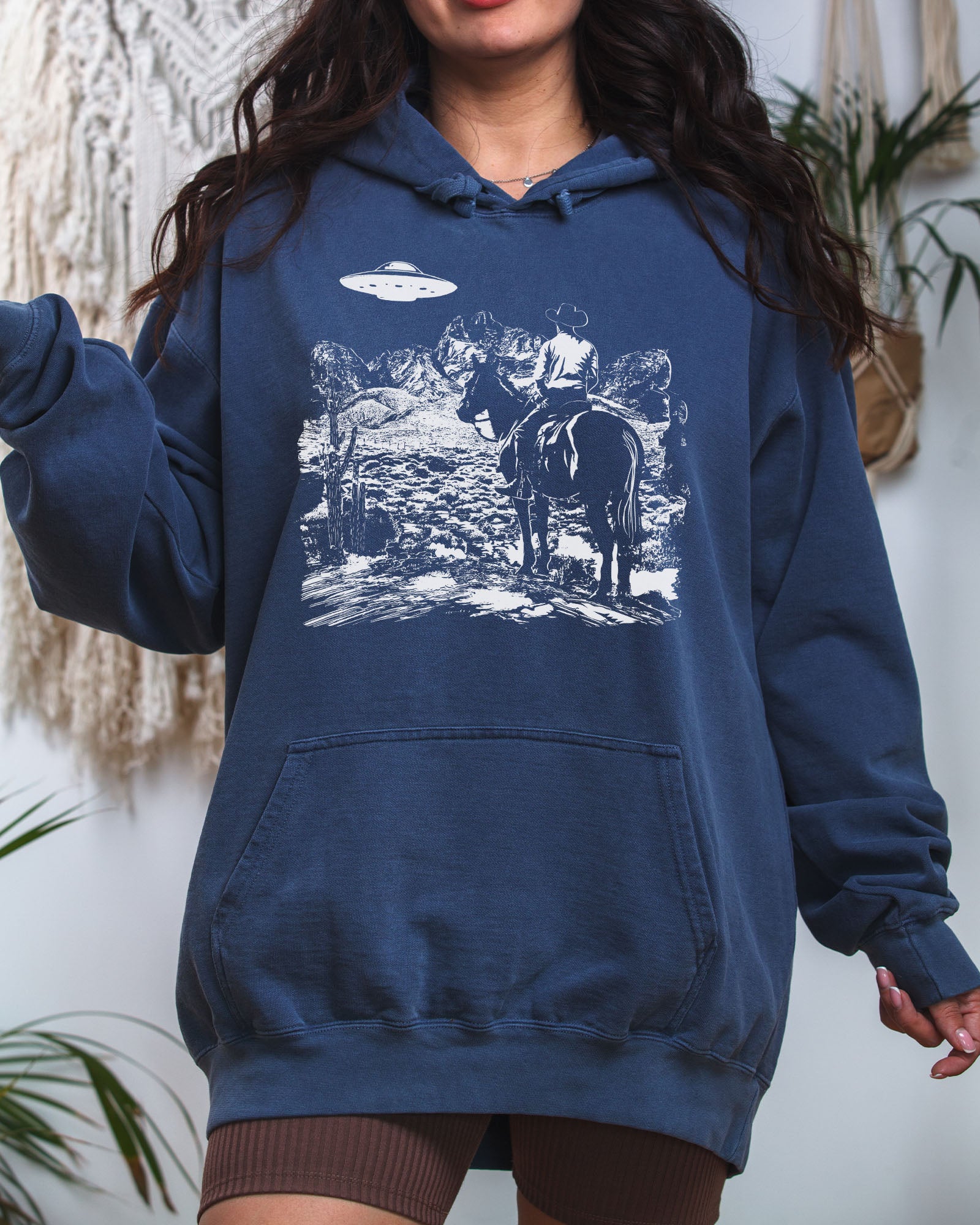 Woman wearing a denim-colored "Cowboy UFO Encounter" hoodie with a retro graphic of a cowboy riding through a desert as a UFO hovers above.