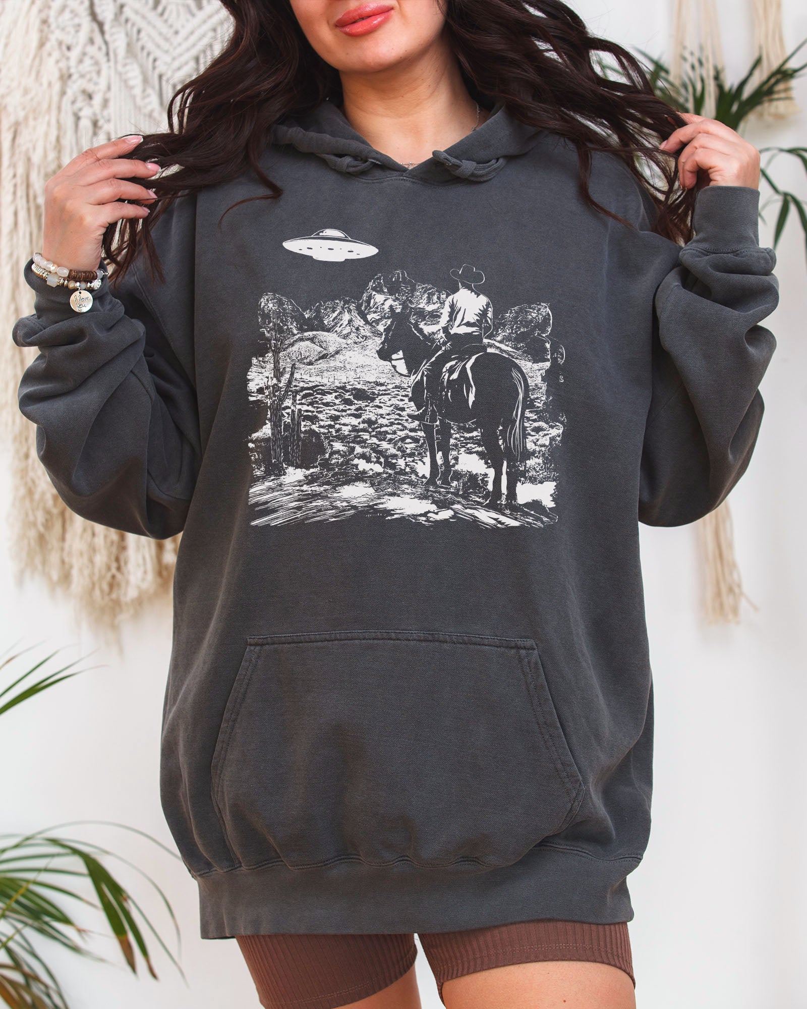 Woman wearing a pepper-colored "Cowboy UFO Encounter" hoodie with a retro graphic of a cowboy riding through a desert as a UFO hovers above.