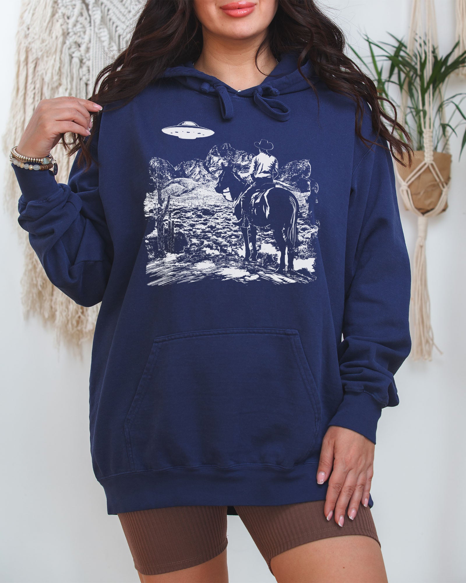 Woman wearing a true-navy "Cowboy UFO Encounter" hoodie with a retro graphic of a cowboy riding through a desert as a UFO hovers above.