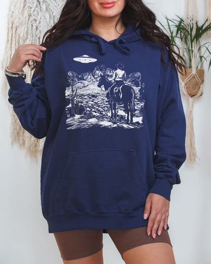 Woman wearing a true-navy "Cowboy UFO Encounter" hoodie with a retro graphic of a cowboy riding through a desert as a UFO hovers above.