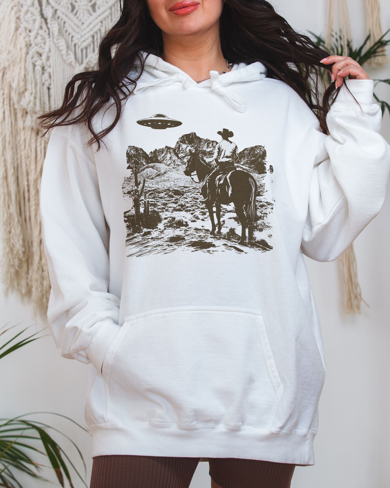 Woman wearing a white-colored "Cowboy UFO Encounter" hoodie with a retro graphic of a cowboy riding through a desert as a UFO hovers above.