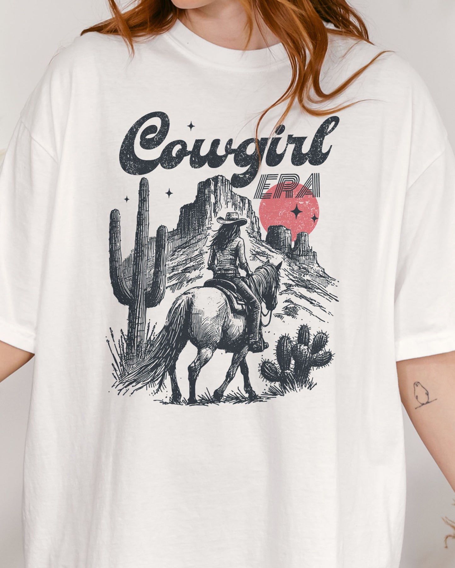 Close-up of woman wearing a white "Cowgirl Era" shirt with a retro graphic of a cowgirl riding a horse through desert scenery.