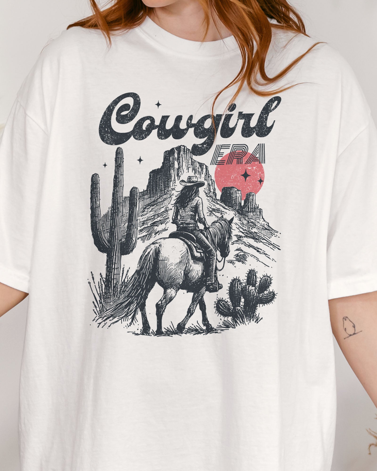 Close-up of woman wearing a white "Cowgirl Era" shirt with a retro graphic of a cowgirl riding a horse through desert scenery.