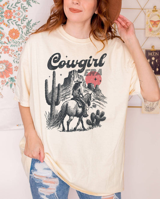 Woman wearing an ivory "Cowgirl Era" shirt with a retro graphic of a cowgirl riding a horse through desert scenery.