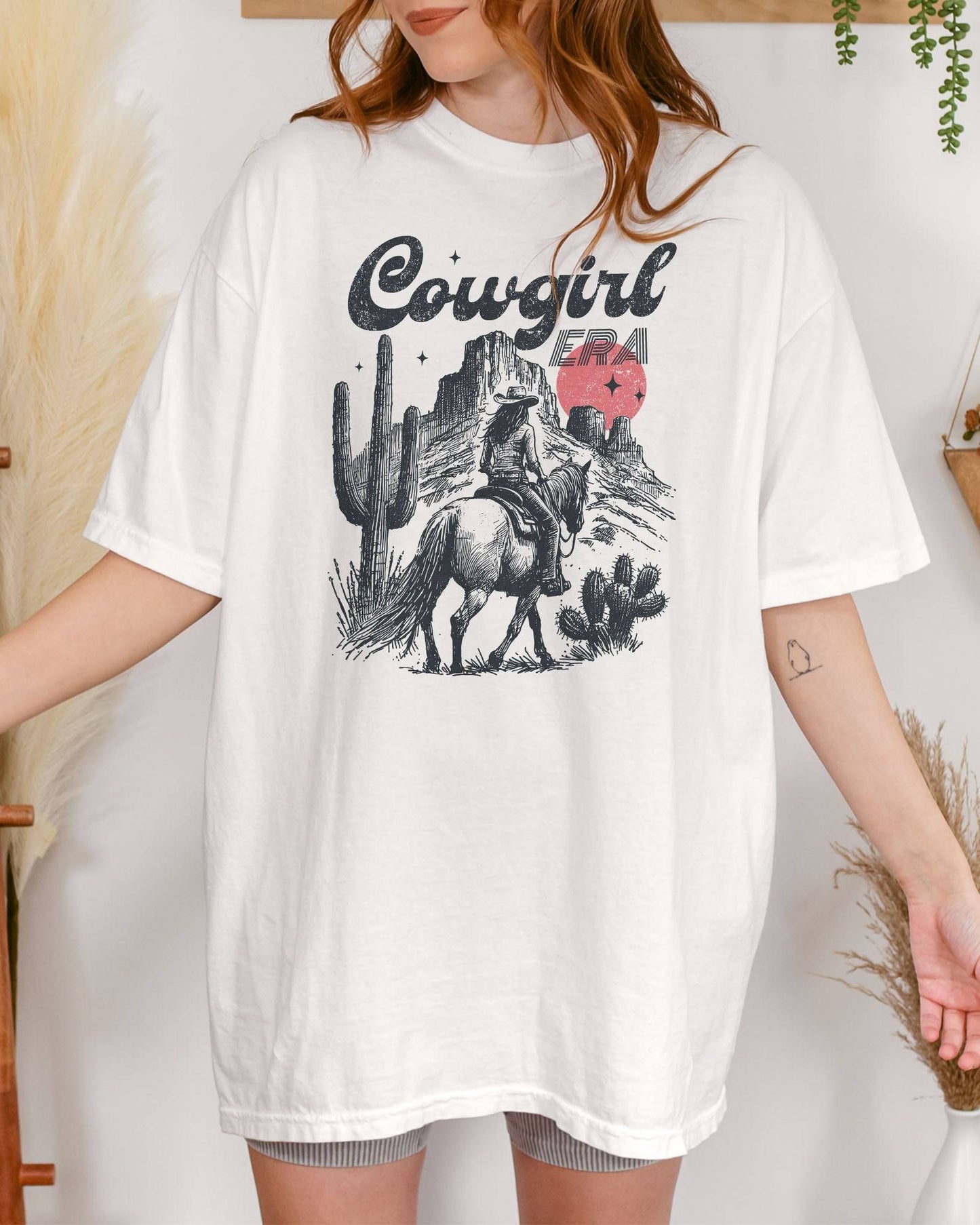 Woman wearing a white "Cowgirl Era" shirt with a retro graphic of a cowgirl riding a horse through desert scenery.