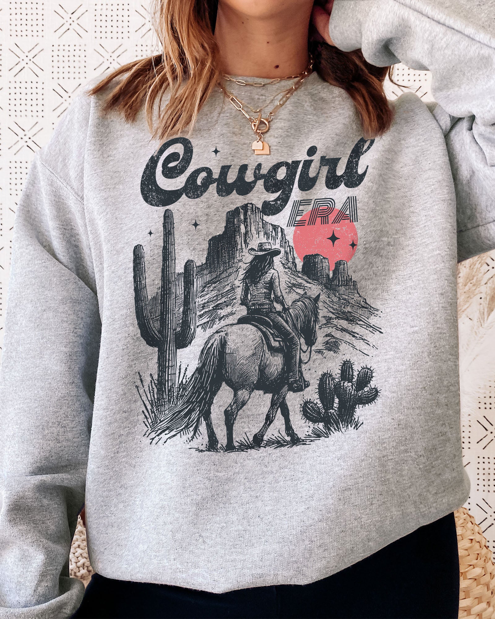 Woman wearing an ash gray sweatshirt featuring a retro cowgirl riding a horse through a Western desert with the lettering 'Cowgirl Era'.