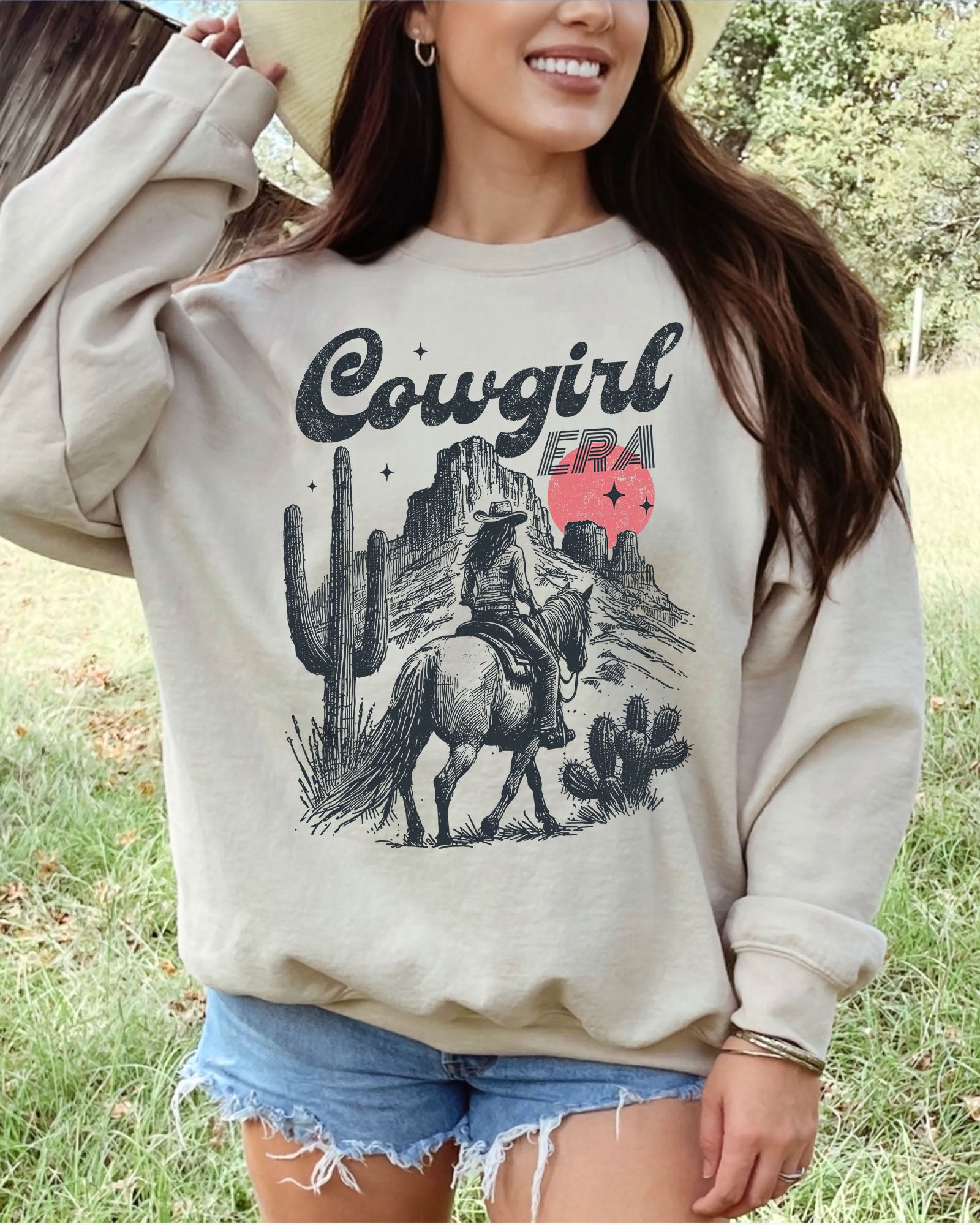 Woman wearing a sand beige sweatshirt featuring a retro cowgirl riding a horse through a Western desert with the lettering 'Cowgirl Era'.