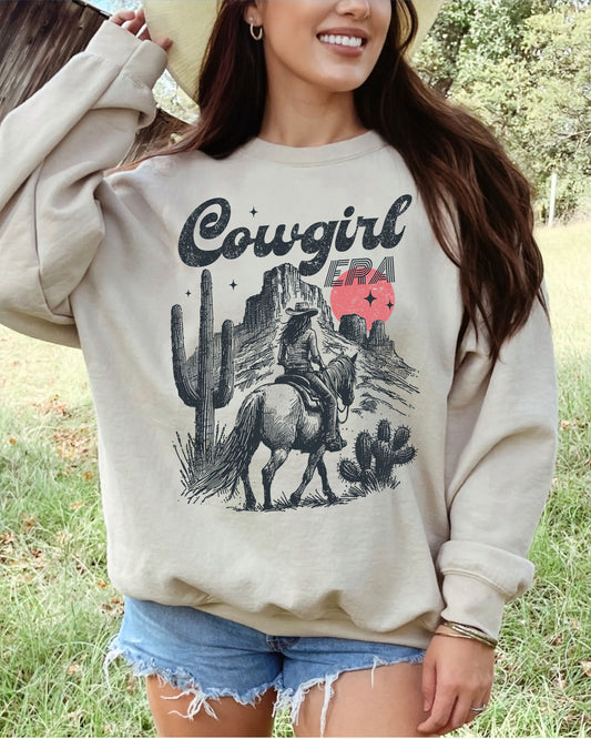 Woman wearing a sand beige sweatshirt featuring a retro cowgirl riding a horse through a Western desert with the lettering 'Cowgirl Era'.