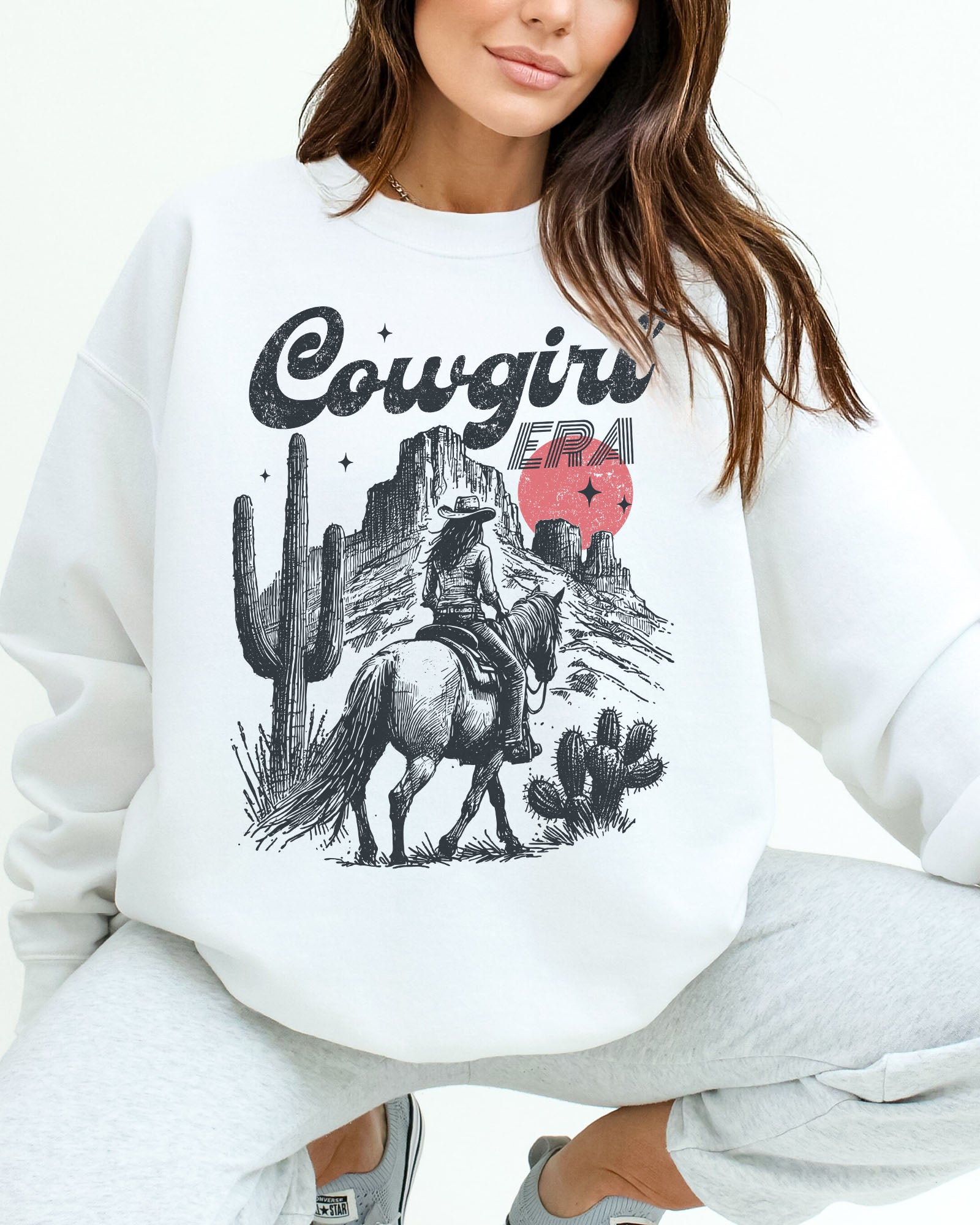 Woman wearing a white sweatshirt featuring a retro cowgirl riding a horse through a Western desert with the lettering 'Cowgirl Era'.