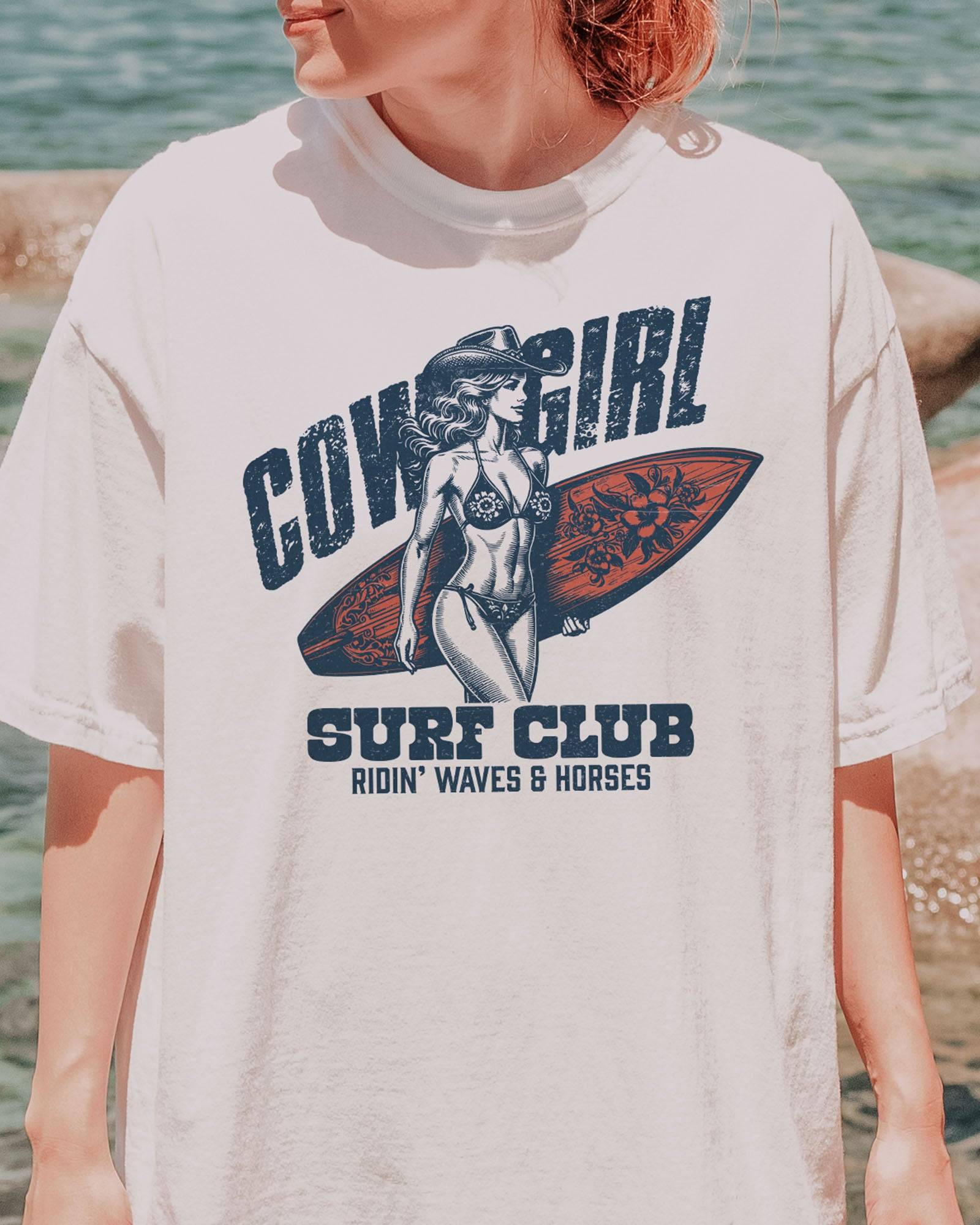 Close-up of woman wearing a white graphic tee at the beach with the quote "Cowgirl Surf Club" and a retro graphic of a cowgirl in a bikini with a surfboard in her hand.