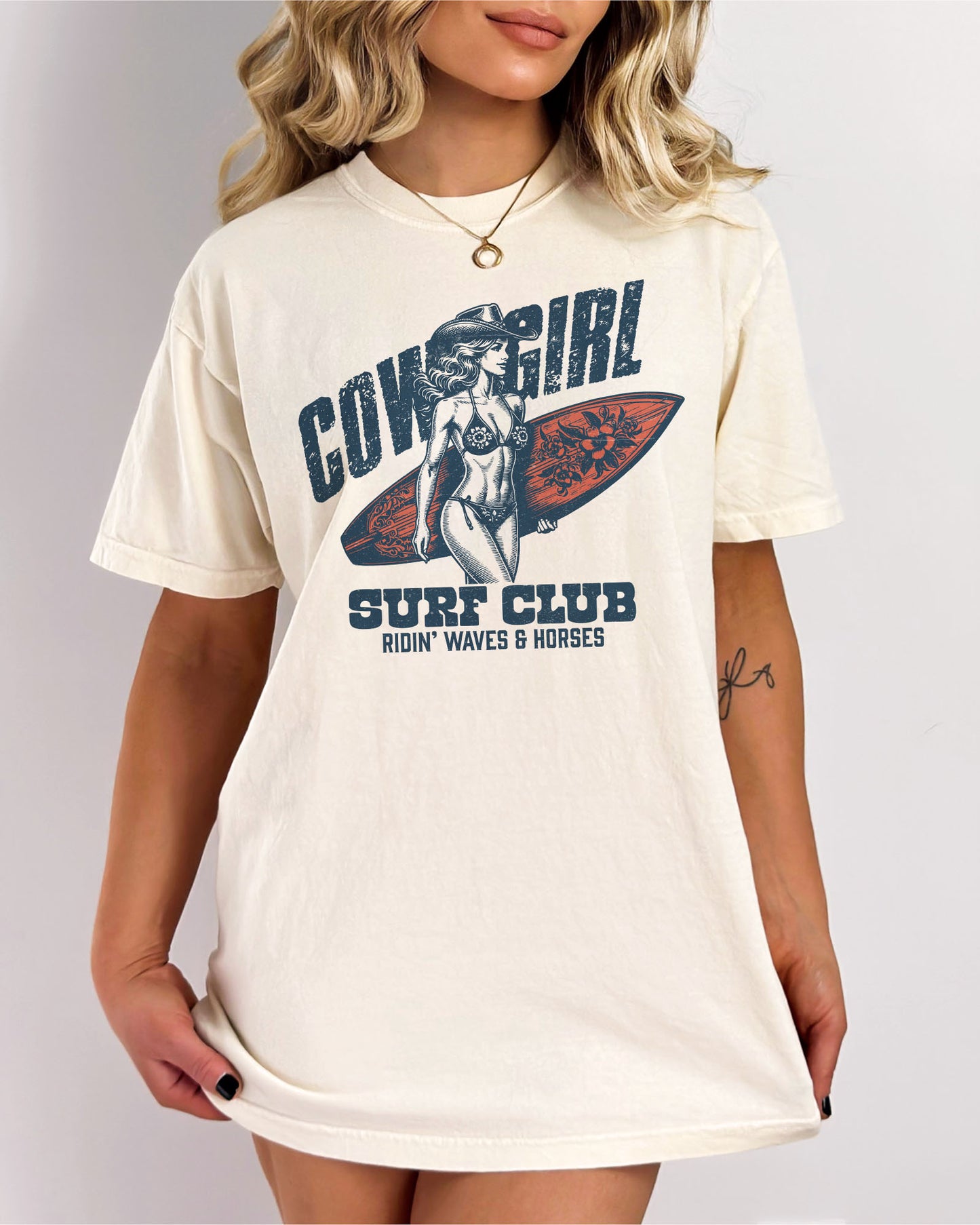 Woman wearing an ivory graphic tee at the beach with the quote "Cowgirl Surf Club" and a retro graphic of a cowgirl in a bikini with a surfboard in her hand.