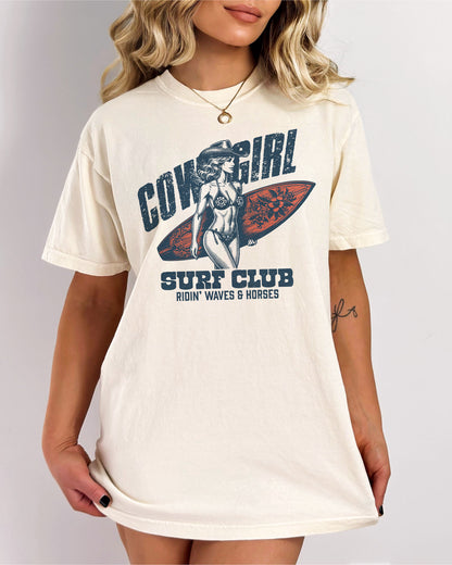 Woman wearing an ivory graphic tee at the beach with the quote "Cowgirl Surf Club" and a retro graphic of a cowgirl in a bikini with a surfboard in her hand.