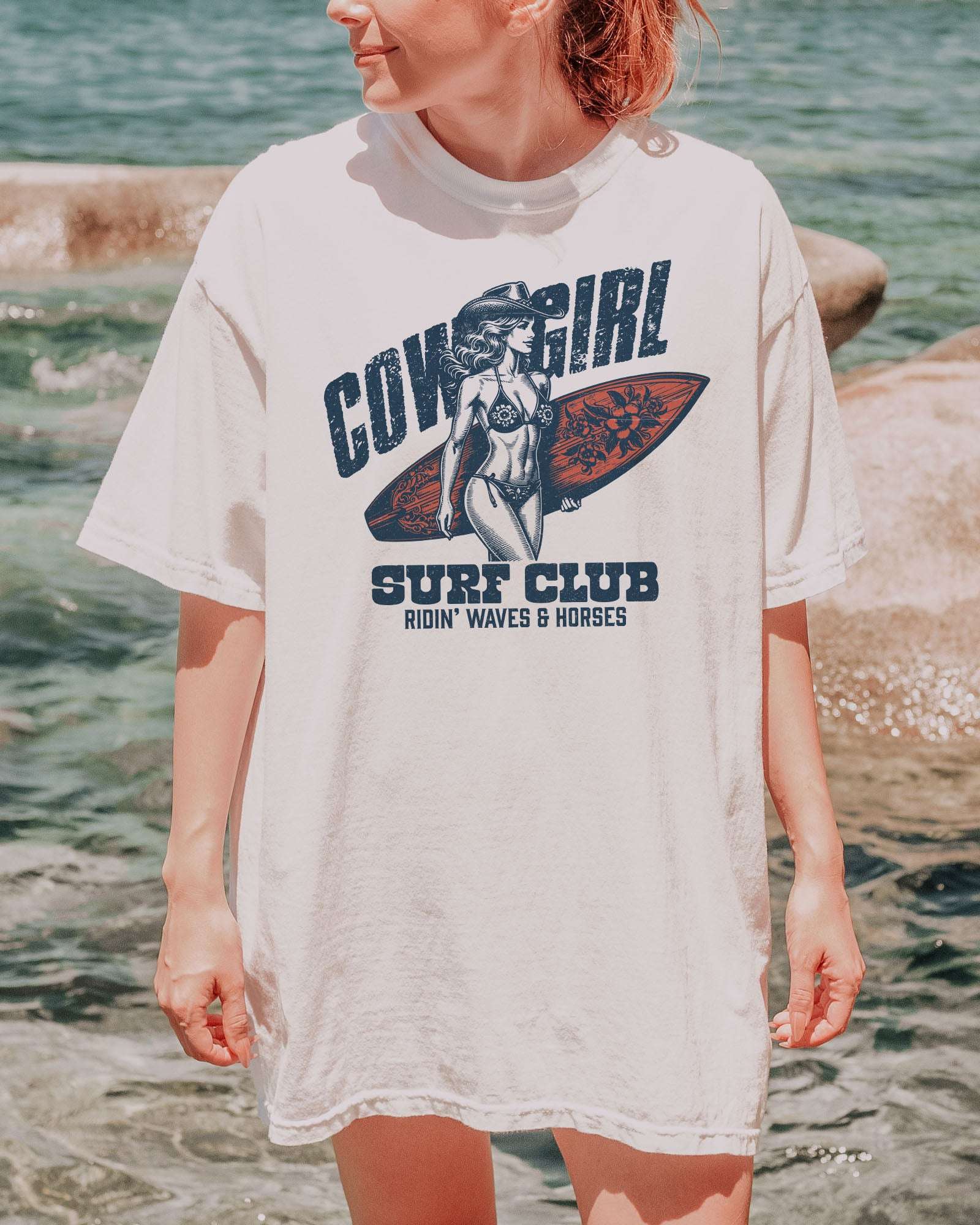 Woman wearing a white graphic tee at the beach with the quote "Cowgirl Surf Club" and a retro graphic of a cowgirl in a bikini with a surfboard in her hand.