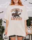 Woman wearing an ivory "Creepin It Country" shirt with a retro Halloween graphic of a pumpkin-headed cowboy riding a horse.
