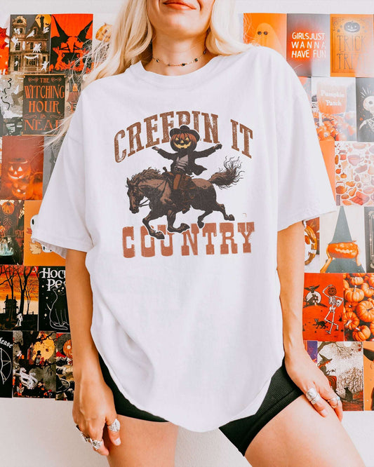 Woman wearing a white "Creepin It Country" shirt with a retro Halloween graphic of a pumpkin-headed cowboy riding a horse.