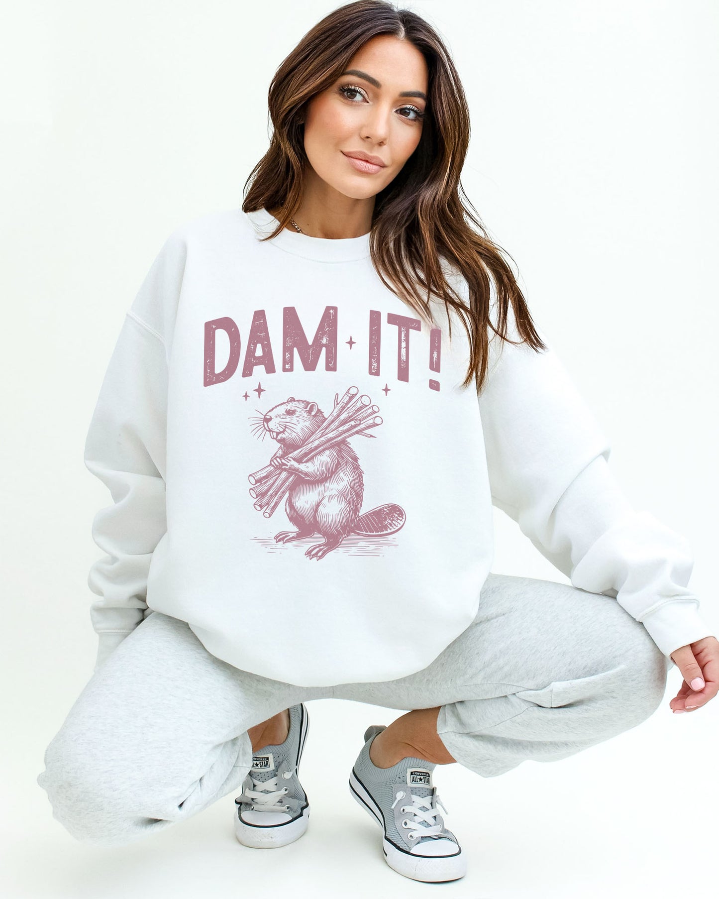 Woman wearing a white “Dam It” sweatshirt with a retro-style beaver graphic and funny expression.