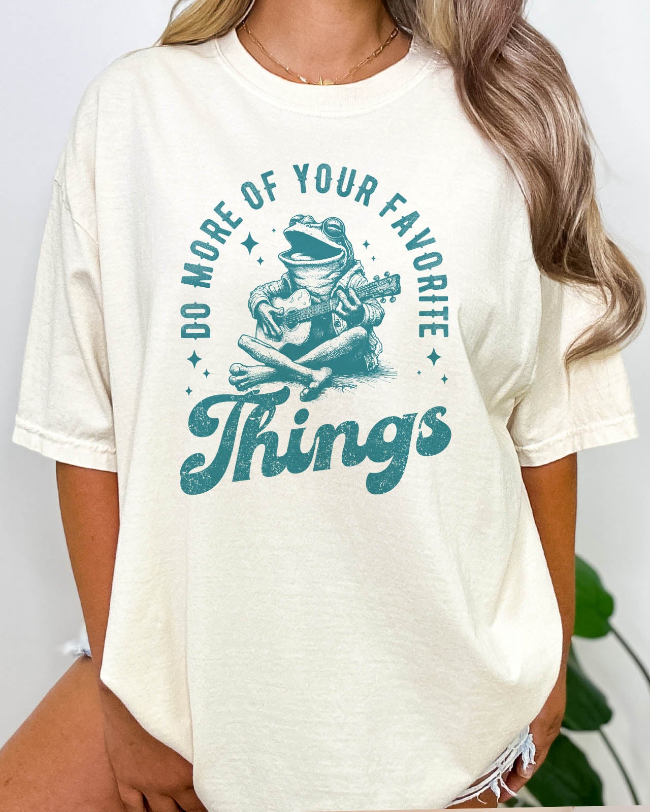 Woman wearing "Do More of Your Favorite Things" shirt in natural beige color with a funny retro frog playing the guitar graphic