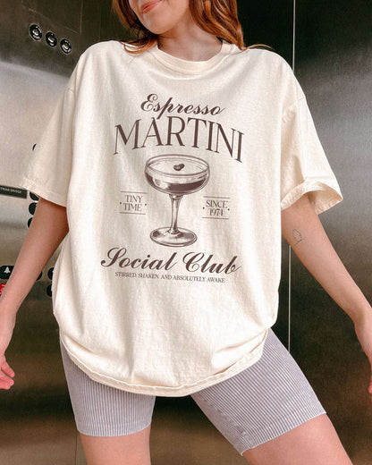 Woman wearing an ivory-colored 'Espresso Martini Social Club – Stirred, Shaken, and Awake' shirt with a retro graphic of an espresso martini glass.