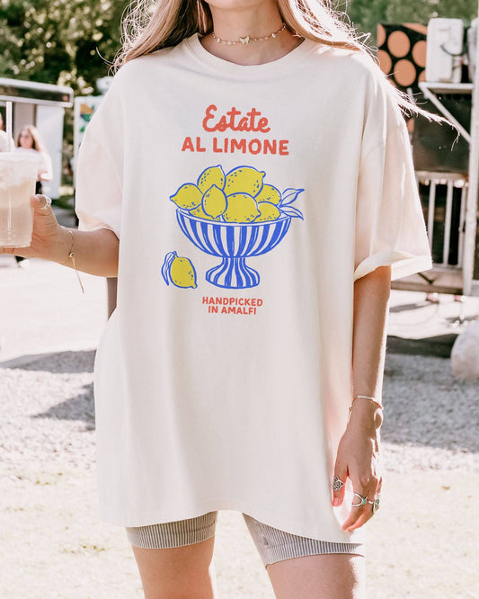 Woman wearing an ivory “Estate Al Limone - Handpicked in Amalfi” shirt with a retro graphic of a fruit bowl filled with lemons.