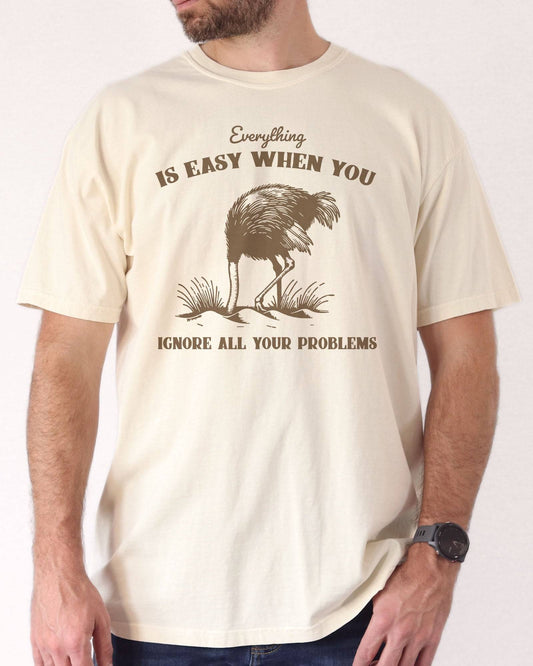 Man wearing ivory color shirt with quote "Everything Is Easy When You Ignore All Your Problems" and a retro graphic of a strauss burying his head in the sand.