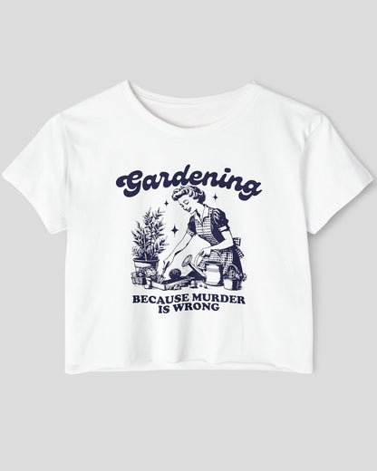 Flat-lay of a white crop top featuring a funny retro-style graphic of a housewife gardening with the text ‘Gardening, Because Murder Is Wrong’.