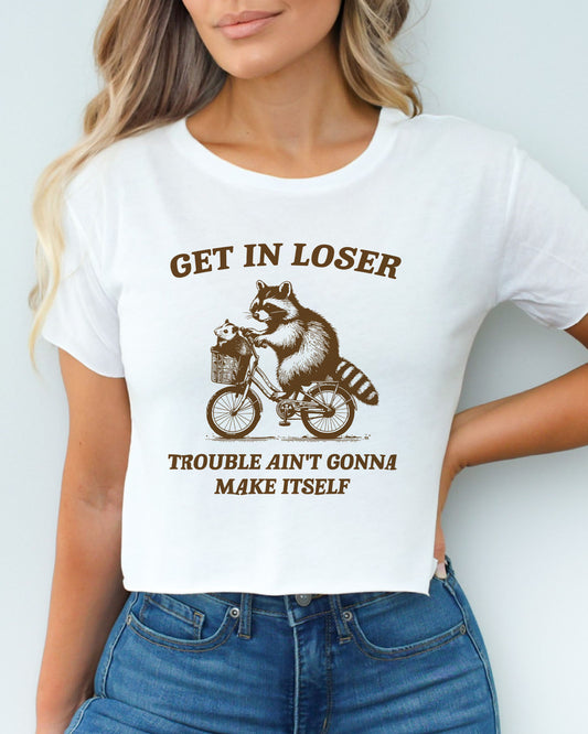 Woman wearing white 'Get In Loser, Trouble Ain't Gonna Make Itself' crop top featuring a funny retro raccoon riding a bicycle.
