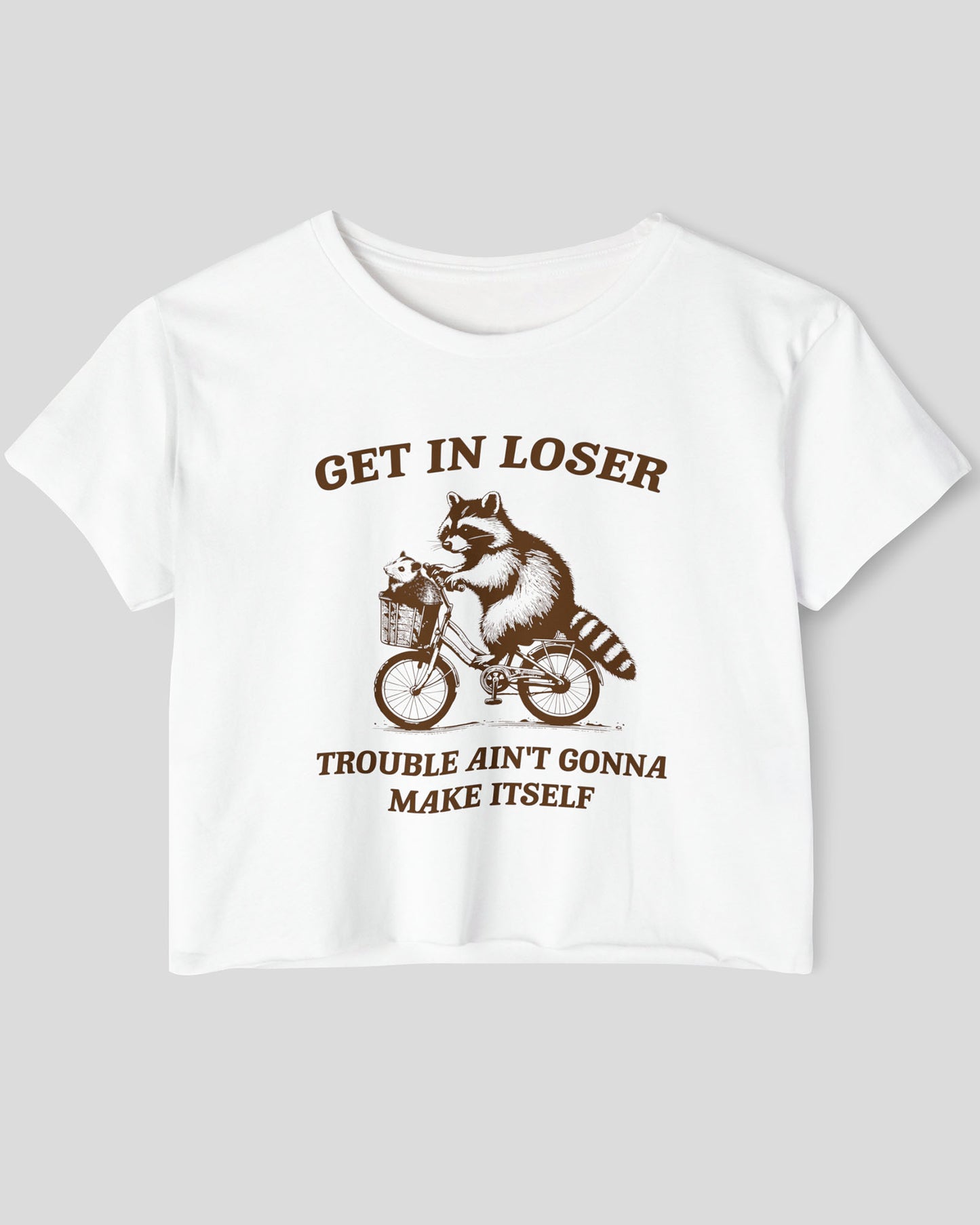 Flat lay white 'Get In Loser, Trouble Ain't Gonna Make Itself' crop top featuring a funny retro raccoon riding a bicycle.