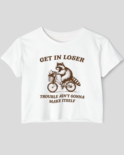 Flat lay white 'Get In Loser, Trouble Ain't Gonna Make Itself' crop top featuring a funny retro raccoon riding a bicycle.