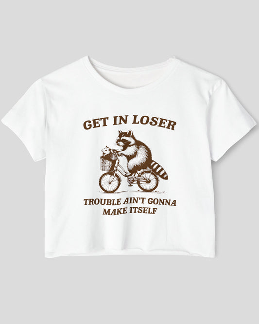 Flat lay white 'Get In Loser, Trouble Ain't Gonna Make Itself' crop top featuring a funny retro raccoon riding a bicycle.