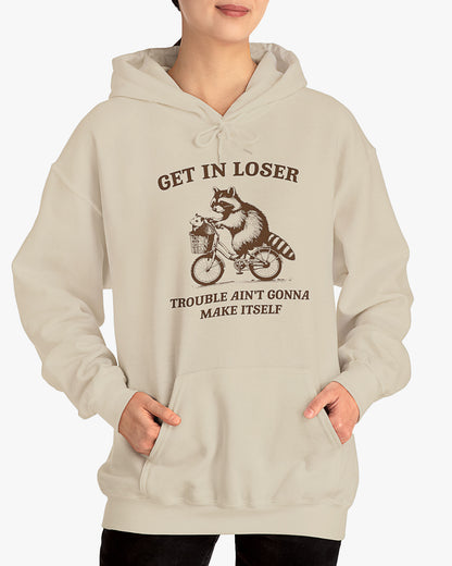 Woman wearing sand beige color hoodie with the quote 'Get In Loser, Trouble Ain't Gonna Make Itself' and a funny retro raccoon riding a bycicle graphic